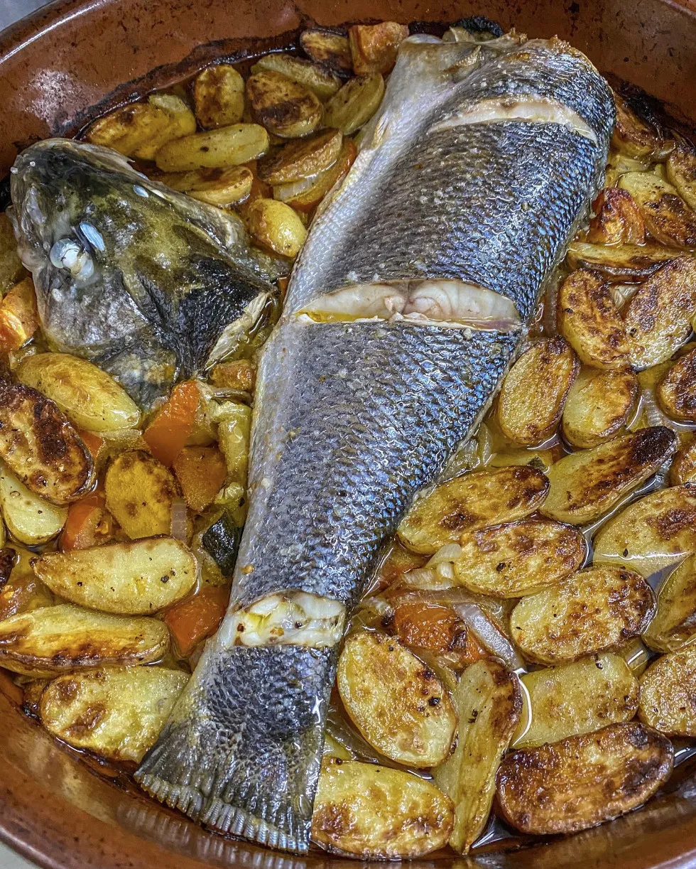 Robalo no forno com batata e legumes, num tabuleiro de barro
